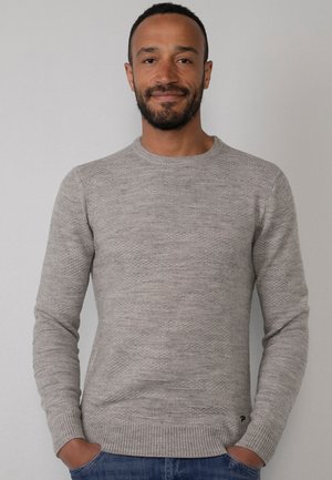 Suéter gris claro con un patrón de punto texturizado, cuello redondo y puños de canalé. Llevado con jeans azules, con las manos en los bolsillos.