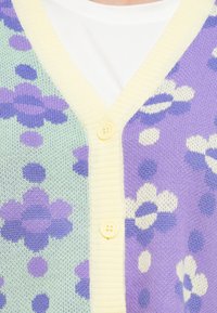 Πλεκτή ζακέτα με παστέλ πράσινο και μωβ λουλουδάτο σχέδιο, με κίτρινο ριμπ λαιμόκοψη και τρία κίτρινα κουμπιά στο μπροστινό μέρος.