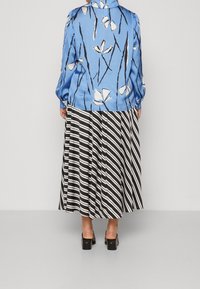 Donna con una camicia a maniche lunghe floreale blu, una gonna plissettata a righe bianche e nere e scarpe con tacco quadrato nere, in piedi contro un muro grigio.