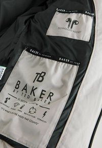Grå indre foring af en jakke med to mærker, et med "TEKNOLOGI LOMME" og det andet med plejeinstruktioner og symboler.