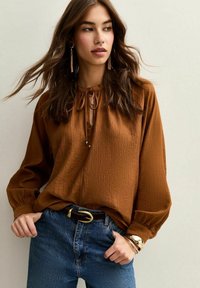 Blouse marron texturé avec un col noué, des manches bouffantes et une coupe ample, associée à un jean bleu taille haute et une ceinture.