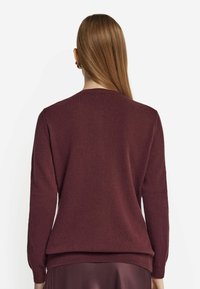 Burgundy stickad tröja med rund halsringning, långa ärmar och en mjuk textur. Har en ribbad nederkant och muddar för ytterligare detaljer.