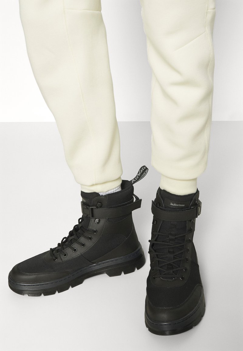 Person, der schwarze Schnür-Ankle Boots mit Riemen und cremefarbenen Hosen trägt, vor einem schlichten Hintergrund.