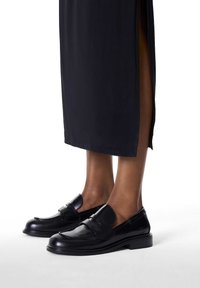 Schwarze Leder-Loafer mit abgerundetem Zehenbereich, dekorativem Riemen vorne, leichtem Absatz und glatter Textur, getragen zu einem langen schwarzen Rock.