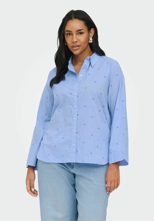Vrouw draagt lichtblauw overhemd met kleine oogjespatronen en lichtblauwe spijkerbroek, poseert tegen een witte achtergrond.
