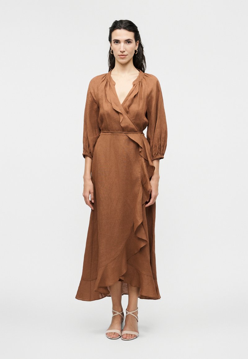 Γυναίκα που φοράει ένα μακρυμάνικο καφέ φόρεμα με βολάν και κρεμ σανδάλια με λουράκια, στέκεται μπροστά σε απλό φωτεινό φόντο.