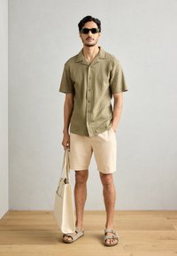Kortærmet olivengrøn tekstureret skjorte, lyse beige shorts, beige tote taske og brune sandaler med spænder, sat mod en neutral baggrund.