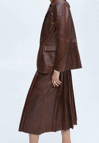 Ruskea nahkatakki, joka on yhdistetty samankaltaiseen laskostettuun nahkahameeseen. Ominaisuuksiin kuuluvat muotoiltu malli ja sileä pinta.