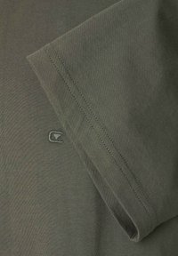 Närbild av olivgrön tyg med dubbelstygna söm och liten broderad logotypdetalj på den släta textilytan.