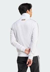 White athletic jacket featuring a ribbed collar and cuffs, side stripes, and a logo accent at the back neck, designed for a fitted silhouette.