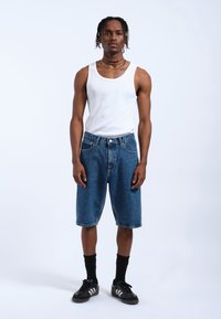 Shorts de mezclilla azules con un ajuste holgado, combinados con una camiseta sin mangas blanca y calcetines negros. El modelo está de pie frente a un fondo blanco liso.