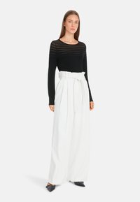 Schwarzes Langarmoberteil mit transparenter gestreifter Gestaltung, kombiniert mit hochgeschnittenen weißen weiten Hosen, die mit einem gebundenen Gürteldetail versehen sind. Schwarze spitze Schuhe.