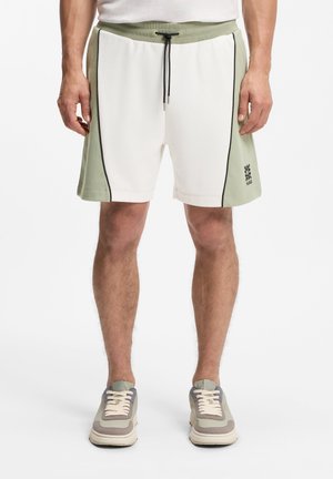 Uomo che indossa pantaloncini con coulisse bianchi e verde oliva con piping nero e sneaker abbinate, in piedi contro uno sfondo bianco.