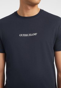 Tamsiai mėlyna medvilninė T-shirt marškiniai su apvalia iškirpte ir trumpomis rankovėmis; krūtinėje išsiuvinėtas „GUESS JEANS“ logotipas balta spalva.