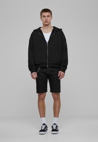 Melna zip-up kapuce ar lielu kapuci, apvienota ar melniem šortiem. Balts bez piedurknēm krekls zem tā, melni un balti sporta apavi, un balti īsie zeķes.