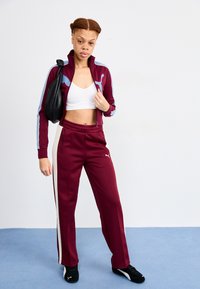 Bordeaux tracksuit met blauwe accenten, aansluitende zip-up jas, bijpassende broeken met zijstrepen, gedragen met een witte crop top en zwarte sneakers.