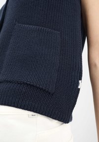Gilet blu navy a maglia con tasca frontale indossato sopra pantaloni bianchi con etichette del marchio visibili su entrambi gli indumenti.