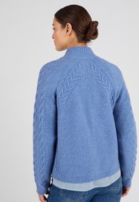 Maglione azzurro chiaro a maglia con colletto alto, motivo a trecce sui maniche e design a costine. Indossato sopra una camicia a righe.