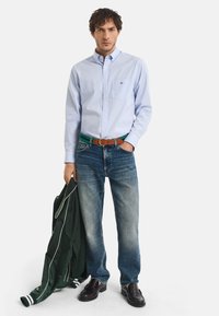 Hellblaues Hemd mit Knöpfen, dunkle Jeans mit Ausbleichungen, brauner Gürtel und schwarze Anzugschuhe. Hält eine grüne Jacke mit weißer Umrandung.