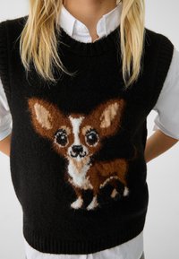 Gilet noir en maille avec un design de Chihuahua duveteux en brun et blanc. Encolure et ourlet côtelés, porté par-dessus une chemise blanche à col.