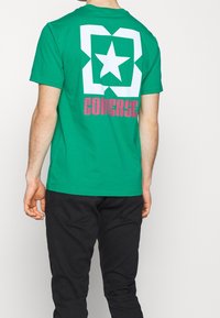 Zelené bavlněné tričko s velkou bílou hvězdou uvnitř čtvercového designu na zadní straně, se slovem "CONVERSE" v růžové pod hvězdou.