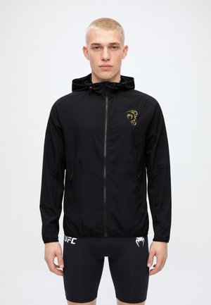 Un modelo masculino está de pie mirando al frente, con una chaqueta negra con capucha y cremallera, un emblema dorado en el pecho y unos pantalones cortos negros de UFC, sobre un fondo blanco liso.