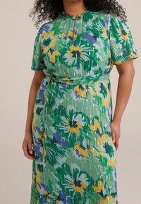 Blommig klänning i grönt med gula och lila accenter. Korta, volangärmar och ett självknutet bälte i midjan. Lätt, texturerad tyg.