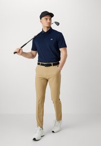 Marinblå pikétröja med korta ärmar, beige chinos, svart bälte och vita sportskor, håller i en golfklubba, bär svart keps.