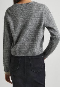 Kobieta w szarym dzianinowym swetrze z prążkowanymi mankietami i pasem, w połączeniu z ciemną spódnicą dżinsową z wysokim stanem i tylnymi kieszeniami.