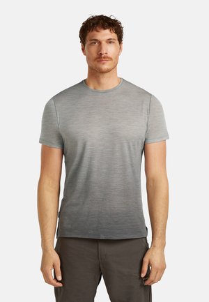 Mężczyzna w szarej koszulce z krótkim rękawem o gradientowym wzorze i ciemnoszarych spodniach, stojący na tle jasnego, jednolitego tła.