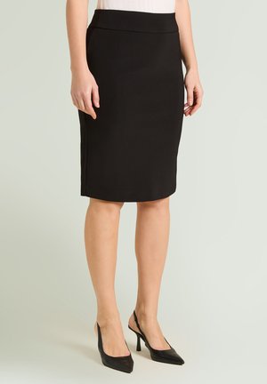Femme portant une jupe crayon noire longueur genou et des talons slingback noirs à bout pointu, debout sur un fond clair.