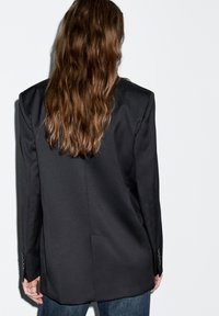 Černé sako s výraznými rameny, hladkou látkou a jedním rozparkem vzadu. Rukávy zdobí knoflíkové detaily. Nositelný přes tmavé džíny.