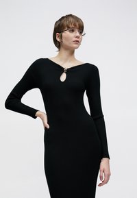 Rochie neagră cu dungi, cu mâneci lungi și decolteu cu cheie. Prezintă un accent auriu la decolteu. Design mulat și textură netedă.