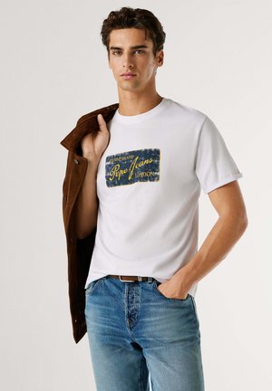 Bela bombažna majica s modrim in rumenim grafičnim tiskom z napisom "Pepe Jeans London", nošena z modrimi jeans hlačami in rjavim semiš jakno.