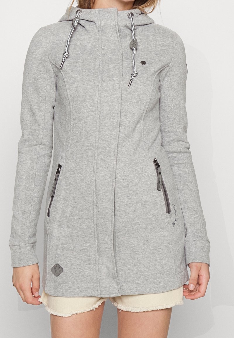 Femme portant une veste à capuche gris clair zippée avec des poches zippées et des cordons, associée à un short de couleur claire.