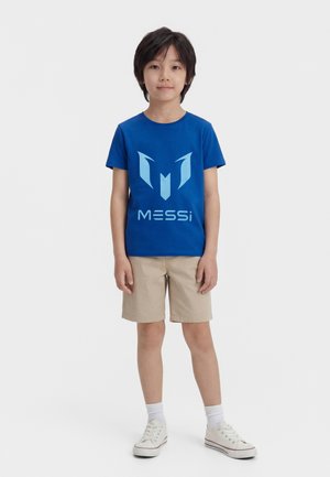 Dziecko stoi przodem, ubrane w niebieską koszulkę z logo Messiego, beżowe szorty, białe skarpetki i białe trampki na jednolitym tle.