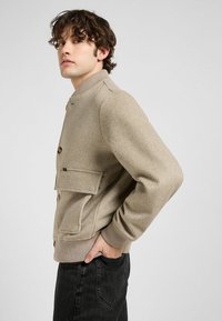 Jonge man met krullend haar draagt een lichtbeige knoopjasje met een voorzak en zwarte jeans, poseert zijwaarts tegen een grijze achtergrond.