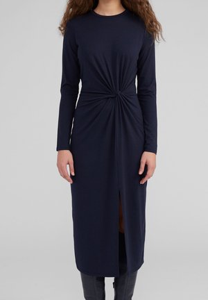 Kobieta w granatowej sukience midi z długim rękawem, z marszczeniem i skręceniem w talii oraz rozcięciem z przodu, w połączeniu z czarnymi kozakami.
