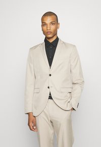 Completo beige con una vestibilità su misura, design monopetto, due tasche frontali, abbinato a una camicia nera e una cintura nera. Tessuto morbido.
