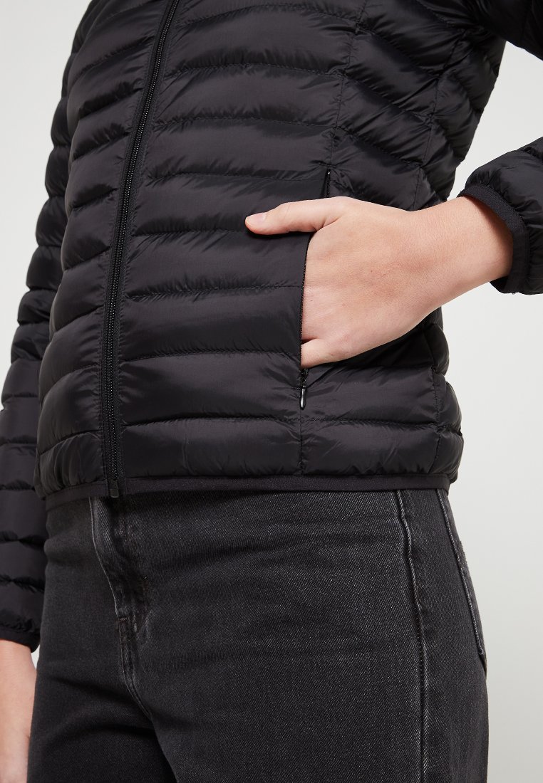 Veste matelassée noire avec fermeture éclair et poches latérales. Elle présente un design matelassé et une texture lisse. Portée sur un jean noir.