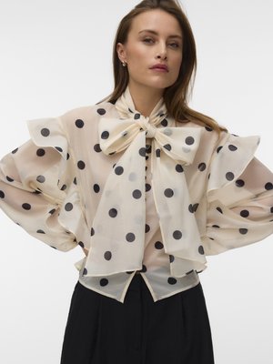 Donna che indossa una camicetta color crema trasparente con grandi pois neri, collo con fiocco oversize e maniche voluminose arricciate, abbinata a pantaloni neri.