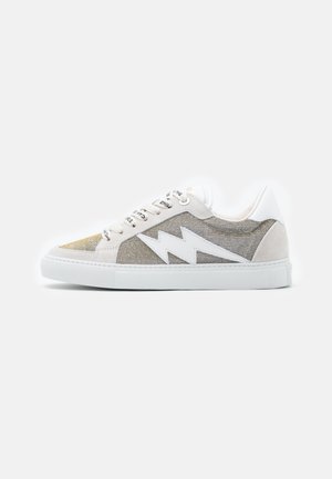 Zapatilla baja con tela beige texturizada, suela blanca, diseño de rayo blanco, detalles en ante blanco y cordones blancos con texto negro.