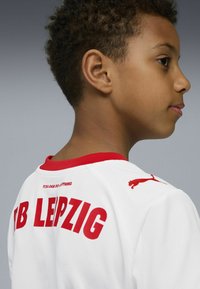 Camiseta deportiva blanca con detalles en rojo. Presenta "RB LEIPZIG" en letras rojas y negrita. Logo de Puma en el hombro. Tela suave y texturizada.