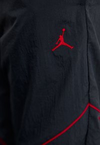 Zwarte stof met een gestructureerd oppervlak bevat een rode geborduurde Jumpman-logo en rode accenten langs de naden.