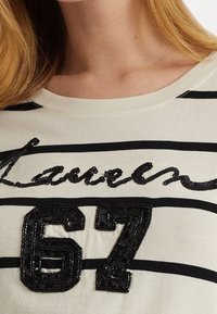 Randig krämfärgad T-shirt med svart paljetttext "Lauren" och paljett "67" på framsidan. Mjuk bomullsmaterial och klassisk rund hals.