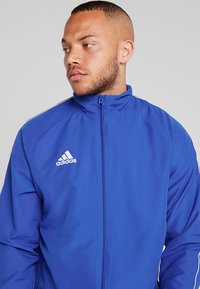 Leichte blaue Jacke mit hohem Kragen, durchgehendem Reißverschluss und weißem Adidas-Logo. Verfügt über kontrastierende weiße Seitenstreifen und eine glatte Textur.