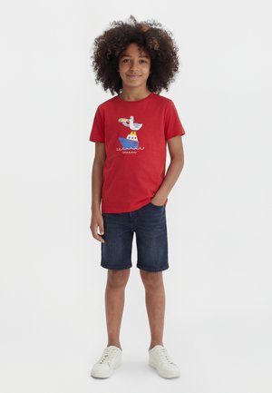 Copil cu păr creț purtând un tricou roșu cu grafică de navă și pasăre, pantaloni scurți din denim închis, pantofi sport albi, stând cu o mână în buzunar.