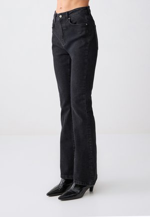 Jean noir taille haute et coupe droite porté par une personne, associé à des bottines noires à bout pointu, sur un fond clair uni.