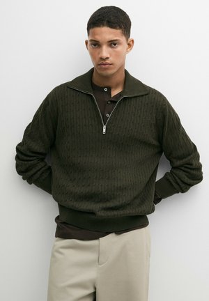 Giovane uomo che indossa un maglione a trecce verde scuro con mezza cerniera sopra una camicia scura e pantaloni beige, in piedi con le mani dietro la schiena.