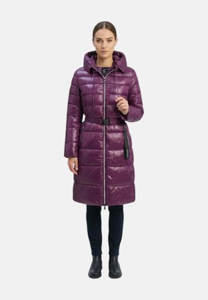 Donna che indossa un lungo cappotto imbottito viola lucido con cappuccio, cintura, pantaloni neri e stivaletti neri, in piedi davanti a uno sfondo semplice.